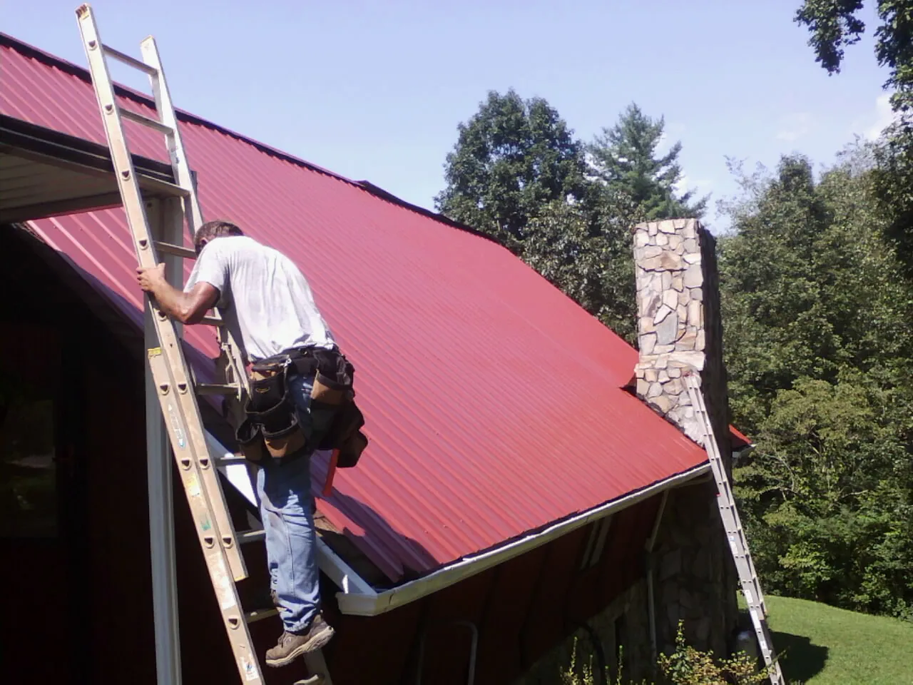 Roofer on a ladder beside a red metal roof and stone chimney — Soffit & Fascia Repair by Roofing Brothers in Drain